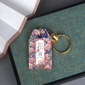 Könnte beinhalten: Ein rosa-blauer Blumen-Schlüsselanhänger mit einer goldenen Glocke und einem Ring. Das Schild trägt japanische Schriftzeichen, die "花見" (hanami) lauten.