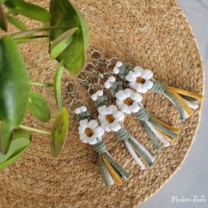 May include: Five crocheted daisy keychains with green and yellow tassels. The keychains are arranged in a circle on a woven straw surface.