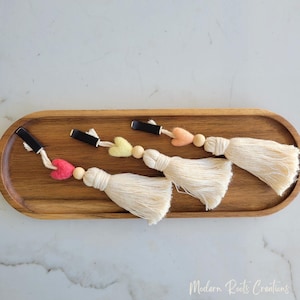 May include: Four cream-colored tassel clips with wooden beads and felt heart accents in pink, yellow, and peach. Each clip has a black metal clasp. The clips are arranged on a wooden oval tray.