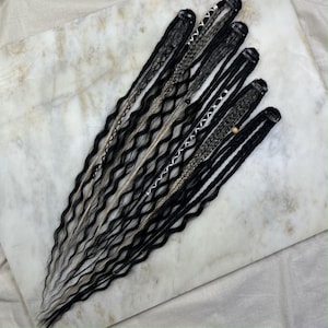 Peut inclure: Un ensemble de dreadlocks synthétiques noires, grises et blanches avec des perles et des clips. Les dreadlocks sont disposées en éventail sur une surface en marbre blanc.