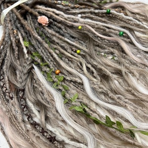 May include: Close-up of a bundle of handmade dreadlocks in shades of gray, brown, and white. The dreads are adorned with small green leaves, beads, and a pink flower. Braided sections add texture to the design.
