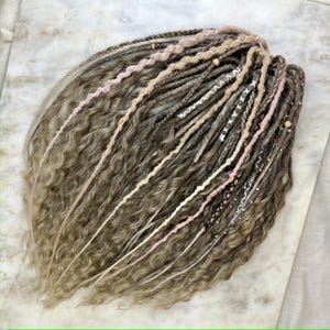 May include: A set of handmade dreadlocks in shades of brown and blonde, with wavy texture. Some dreads are accented with pink, white, and beige yarn wraps, and wooden beads. The dreadlocks are styled in a loose, flowing arrangement.