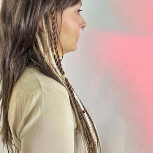 Könnte beinhalten: Eine Person mit langen braunen Haaren mit weißen und braunen Zöpfen. Die Zöpfe haben kleine Perlen und Charms. Die Person trägt ein hellbraunes Shirt.