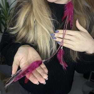 May include: A pink feather hair extension with a braided cord and silver beads. The extension is shown in a person's hand and hanging down from their ear.