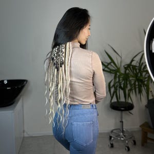 May include: A woman with long black hair with blonde and black dreadlocks, wearing a beige turtleneck shirt and blue jeans.