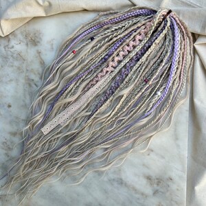 May include: A set of synthetic dreadlocks in shades of blonde and lavender with white and pink accents. The dreadlocks are wavy and have beads and lace accents.