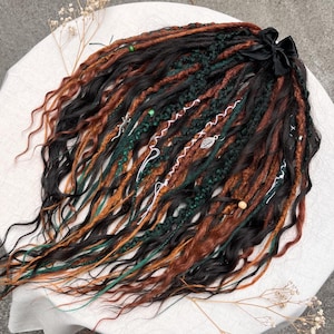 May include: A collection of handmade dreadlocks in shades of black, brown, and green, with decorative accents. The dreadlocks are styled with a black ribbon and various charms, including a leaf and beads. The hair extensions are arranged on a white surface.