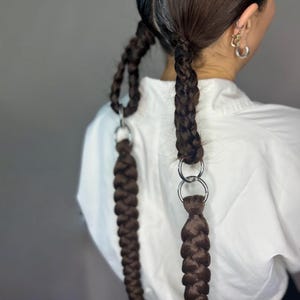 Op de afbeelding: Twee bruine vlechten met zilveren ringen aan de uiteinden. De vlechten zijn aan elkaar gebonden met een zilveren ring.