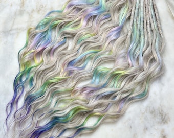 Op de afbeelding: Een verzameling golvende, lichtgrijze synthetische dreadlocks met pastelkleurige regenbooghighlights. De dreadlocks zijn gerangschikt in een vloeiend patroon, met secties in blauw, groen, geel en paars. Een haaraccessoire.
