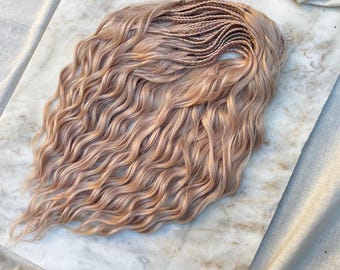 May include: A light brown wig with wavy hair and braided roots. The wig features small braids at the top, transitioning into flowing, voluminous waves. The hair has a natural, sun-kissed colour.