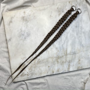 May include: Two long, braided brown hair extensions with silver metal rings at the top. The extensions are lying on a white and grey marble surface. The braids are thick and evenly woven.