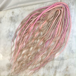 Puede incluir: Un conjunto de extensiones de cabello sintético en una mezcla de tonos rubios y rosas. El cabello está trenzado y ondulado.