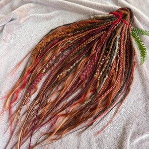May include: A bundle of handmade dreadlocks in shades of red, orange, and brown, tied together with a red ribbon. The dreads feature various textures, including braids and wrapped sections, with gold accents.