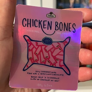 May include: A pink and blue sticker with the text "CHICKEN BONES" and a drawing of a pillow filled with pink and white candy. The text "Spicy CINNAMON candy filled with a Bittersweet CHOCOLATE" is below the drawing. The text "180g 6.3oz" is at the bottom of the sticker.