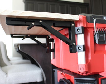 May include: A red toolbox with a wooden work surface attached via black folding brackets. The work surface is light-colored wood. The toolbox has a black handle and a silver latch. The setup appears to be for a mobile workstation.