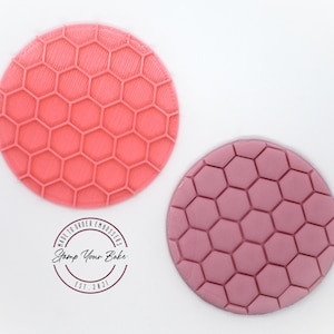May include: Two round silicone embossing stamps with a honeycomb pattern. The stamp on the left is pink and the stamp on the right is a darker shade of pink. The stamps are made by "Stamp Your Bake" and are made to order.