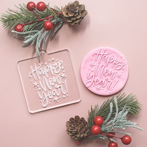 May include: A clear square and a pink circular ornament, both with the white script text "Happy New Year!" and snowflake details. Festive pine branches, red berries, and a pine cone add to the holiday theme.