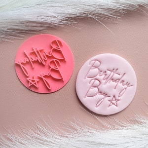 May include: Two pink, circular stamps with cursive text. One stamp reads "Birthday Boy" with a star. The other stamp has the text "You're the best" with a star and heart symbols. The stamps are for cake decorating.