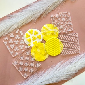 May include: Six clear plastic stencils featuring various honeycomb and bee designs. The stencils are displayed on a light brown surface with white fluffy material.