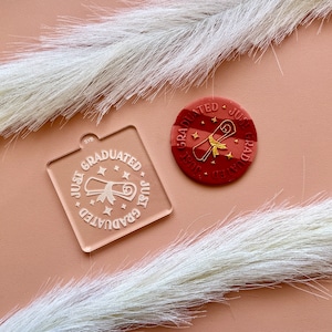 May include: Two acrylic stamps with the text "Just Graduated" and a graduation cap icon. The stamps are white on a clear background. The larger stamp is a circle and the smaller stamp is a square.