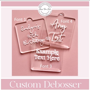 May include: Three clear acrylic debosser stamps with raised white lettering. One stamp reads Evelyn's 30th Birthday, another reads Any Text, and the third reads Example Text Here. Each square stamp features a small tab at the top with SYB printed on it.