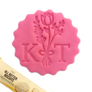 May include: A circular pink scalloped biscuit stamp featuring an embossed floral bouquet design with a tulip and sprigs, flanked by the capital letters K and T. A partial package of M&S Food All Butter Biscuits is visible at the bottom left corner.