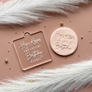 May include: Two clear acrylic stamps with the text "Hugs Kisses and lots of Birthday Wishes" in white lettering. The stamps are on a pink surface with gold glitter.