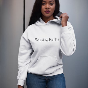Op de afbeelding: Een wit hoodie met de tekst "WALK by FAITH" erop gedrukt. Het model draagt de hoodie met een blauwe jeans.