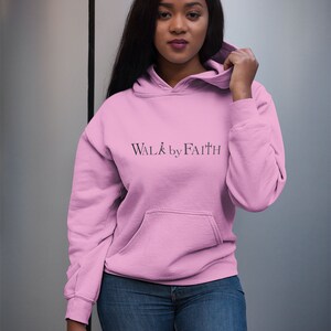 Op de afbeelding: Een roze hoodie met de tekst "WALK by FAITH" in zwarte letters.