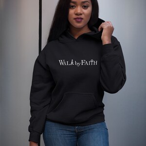 Op de afbeelding: Een zwarte hoodie met een witte afbeelding met de tekst "WALK by FAITH".