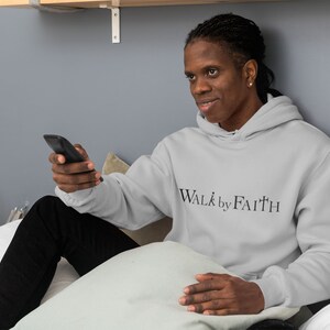 Op de afbeelding: Een persoon draagt een lichtgrijze hoodie met de tekst "WALK by FAITH" in zwarte letters. De persoon zit op een bed en houdt een zwarte afstandsbediening vast.