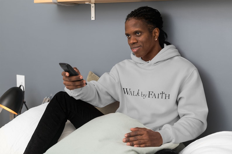 Op de afbeelding: Een persoon draagt een lichtgrijze hoodie met de tekst "WALK by FAITH" in zwarte letters. De persoon zit op een bed en houdt een zwarte afstandsbediening vast.