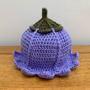 May include: A handmade crochet hat shaped like a purple flower. It has a scalloped edge and a green, leaf-like top. The hat is displayed on a wooden surface.