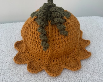 May include: A mustard yellow crochet hat shaped like a pumpkin, featuring a dark olive green stem and curly tendrils at the top. The brim of the hat has a scalloped, petal-like edge, and the main body is constructed with a textured stitch pattern.
