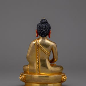 May include: A gold-colored statue of a seated Buddha with a black hair bun and red earlobes. The statue is on a gold-colored base with a lotus design.