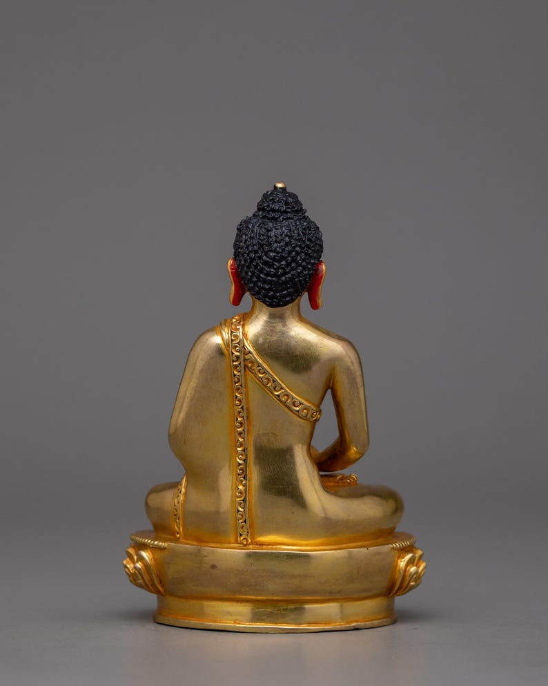 May include: A gold-colored statue of a seated Buddha with a black hair bun and red earlobes. The statue is on a gold-colored base with a lotus design.