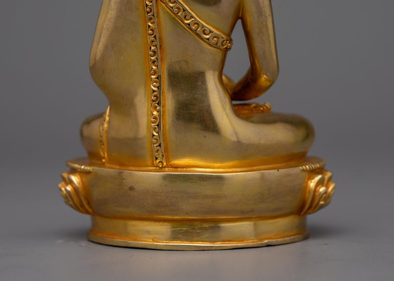 May include: A gold-colored statue of a seated Buddha, with intricate details on the robe and base. The statue is made of metal and has a smooth, polished finish.