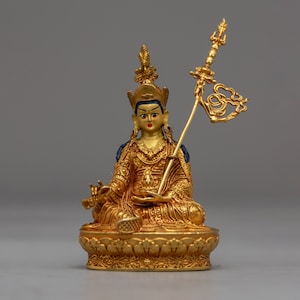 Puede incluir: Una estatua dorada de una figura sentada con una corona y un cetro. La figura lleva una túnica con detalles intrincados y tiene una expresión serena.