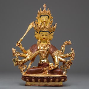 Puede incluir: Estatua dorada de una deidad con múltiples brazos y rostros, adornada con una corona y detalles intrincados. La figura sostiene una espada y otros objetos simbólicos, sentada sobre una base detallada. La estatua tiene una prenda marrón y roja.