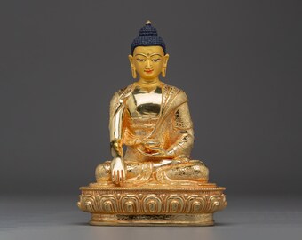 May include: A gold Buddha statue in a seated pose, with a calm expression. The statue has a blue topknot and detailed patterns on the robes and base. The statue is approximately 20 cm tall.