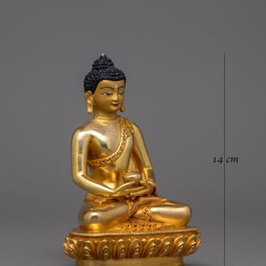 May include: A gold-colored Buddha statue sitting in meditation pose. The statue is 9.5 cm wide and 14 cm tall. The statue is made of metal and has a detailed design.