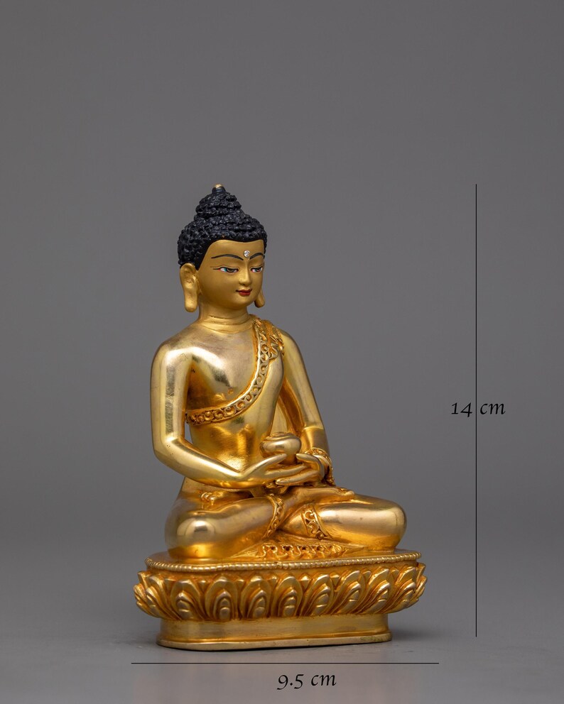 May include: A gold-colored Buddha statue sitting in meditation pose. The statue is 9.5 cm wide and 14 cm tall. The statue is made of metal and has a detailed design.