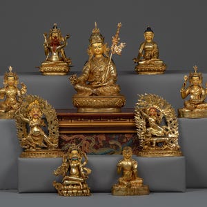 May include: A collection of nine gold-colored statues of deities, some with intricate details and colorful accents, arranged on a tiered platform with a patterned fabric covering.