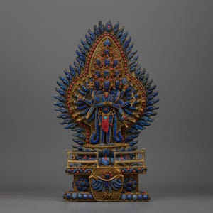 Op de afbeelding: Een gedetailleerde, sierlijke sculptuur met een figuur met meerdere armen in blauw, goud en rood. De figuur is omgeven door een halo van gestileerde vlammen en rust op een decoratieve basis. De sculptuur heeft ingewikkelde details.