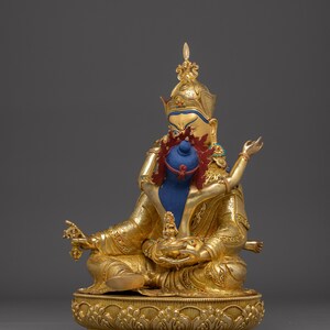 K&ouml;nnte beinhalten: Goldfarbene Statue einer sitzenden Gottheit mit blauem Gesicht und aufwendigem Kopfschmuck. Die Statue h&auml;lt ein blaues Objekt und ist mit roten und goldenen Akzenten verziert. Der Sockel ist mit detaillierten floralen Mustern versehen, vor einem grauen Hintergrund.