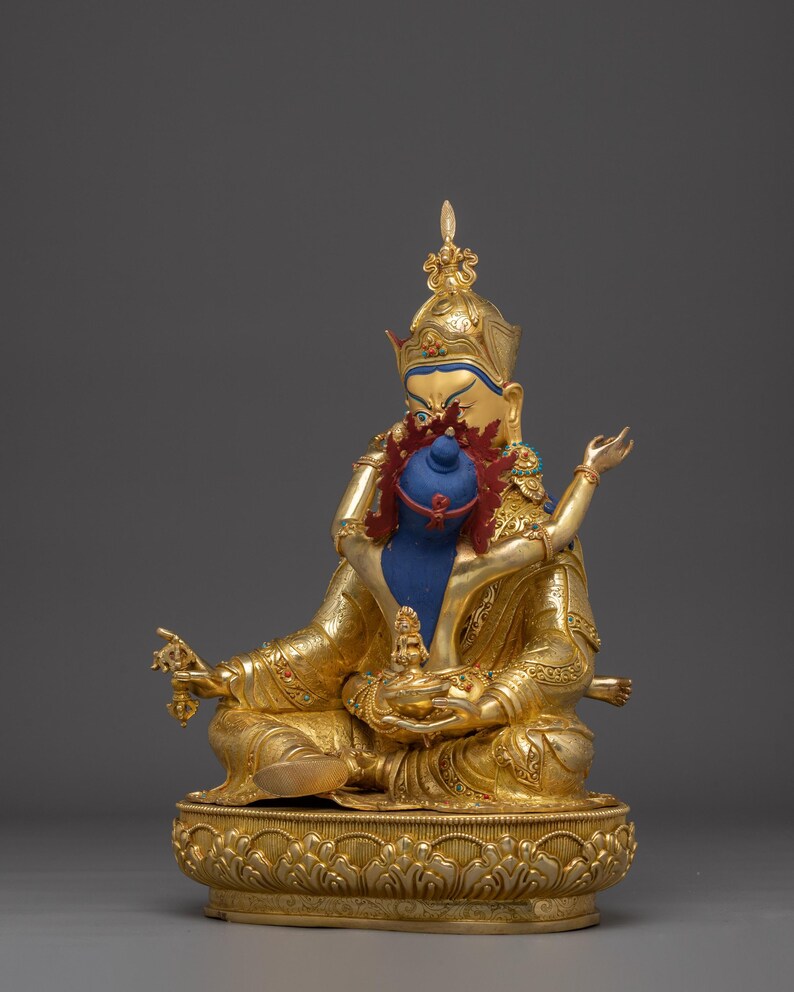 K&ouml;nnte beinhalten: Goldfarbene Statue einer sitzenden Gottheit mit blauem Gesicht und aufwendigem Kopfschmuck. Die Statue h&auml;lt ein blaues Objekt und ist mit roten und goldenen Akzenten verziert. Der Sockel ist mit detaillierten floralen Mustern versehen, vor einem grauen Hintergrund.