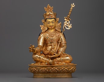 May include: A gold-colored statue of a deity sitting in a meditative pose. The statue is adorned with intricate details and colorful gemstones. The deity is holding a staff in their right hand and a lotus flower in their left hand.