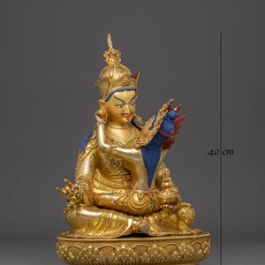 K&ouml;nnte beinhalten: Eine goldene Statue einer sitzenden Figur, geschm&uuml;ckt mit einer Krone und aufwendigen Details. Die Figur h&auml;lt ein blaues und rotes Objekt. Die Statue ist etwa 40 cm hoch und 27 cm breit und ruht auf einem verzierten Sockel.