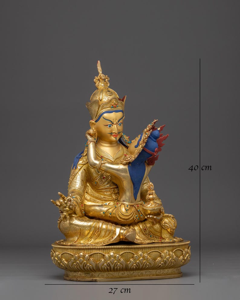 K&ouml;nnte beinhalten: Eine goldene Statue einer sitzenden Figur, geschm&uuml;ckt mit einer Krone und aufwendigen Details. Die Figur h&auml;lt ein blaues und rotes Objekt. Die Statue ist etwa 40 cm hoch und 27 cm breit und ruht auf einem verzierten Sockel.