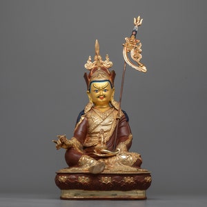 May include: A gold and red statue of a seated figure with a crown and a scepter. The figure has a yellow face with blue eyes and a red mouth. The statue is sitting on a lotus base.